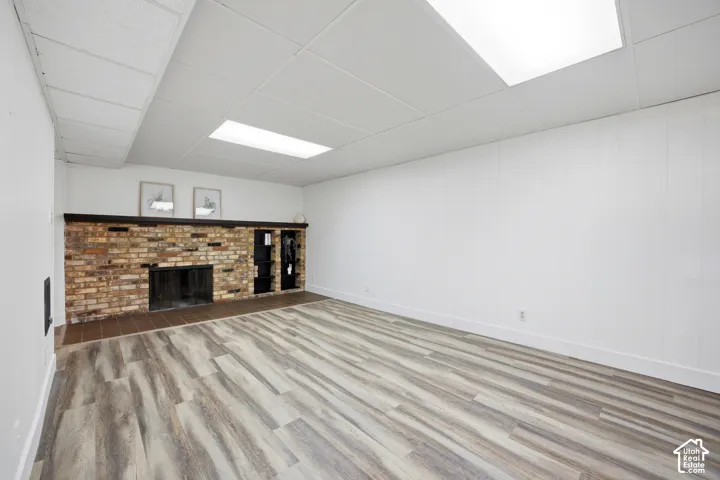 Wood-finish flooring, brick fireplace with a dark wood mantel, built-in shelving, white paneled walls, and drop ceiling with integrated lighting