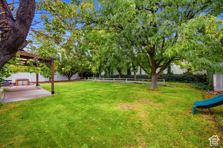 Expansive backyard featuring a lush lawn, mature trees, a concrete patio, a wood pergola, and a white privacy fence