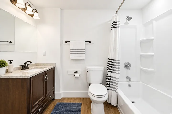 Wood-finish flooring, dark wood vanity with white rectangular sink, light-toned countertop, and a white bathtub with integrated shelving