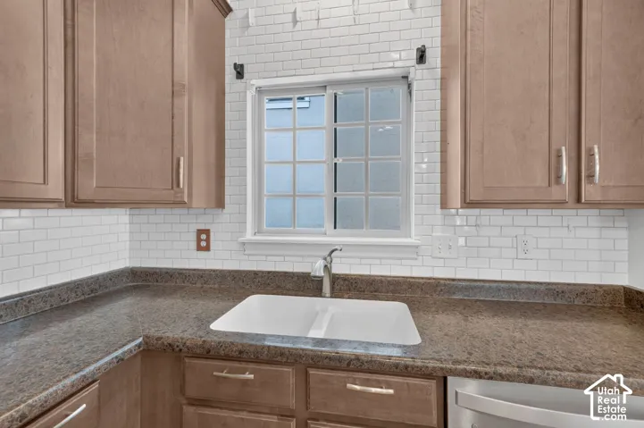 Light wood-finish cabinetry with gold-tone hardware, white subway tile backsplash, dark countertop with an undermount sink, and a white framed window