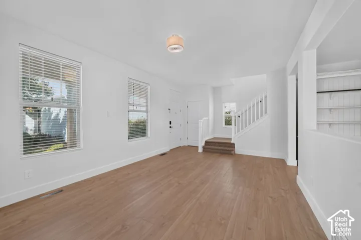 Wood-finish flooring, white walls, two windows with blinds, a staircase with white balusters, and an entryway with a light fixture