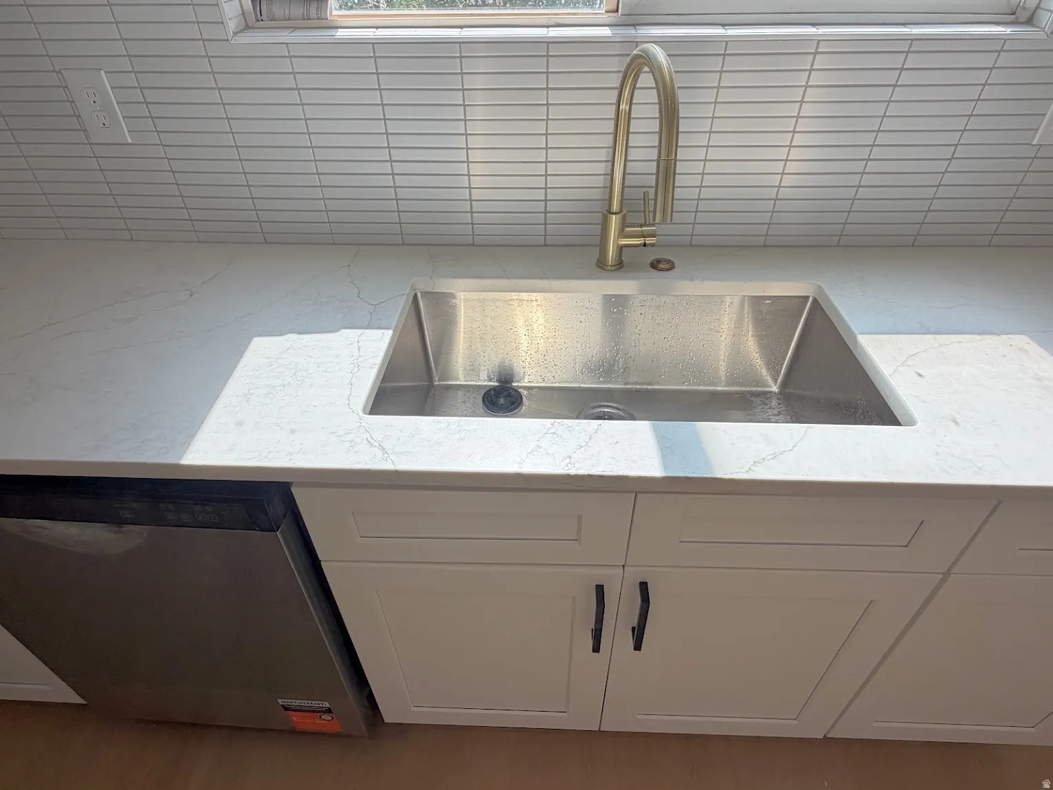 White shaker-style cabinetry with black hardware, stainless steel sink, gold-finish gooseneck faucet, light-toned countertop with veining, and linear tile backsplash