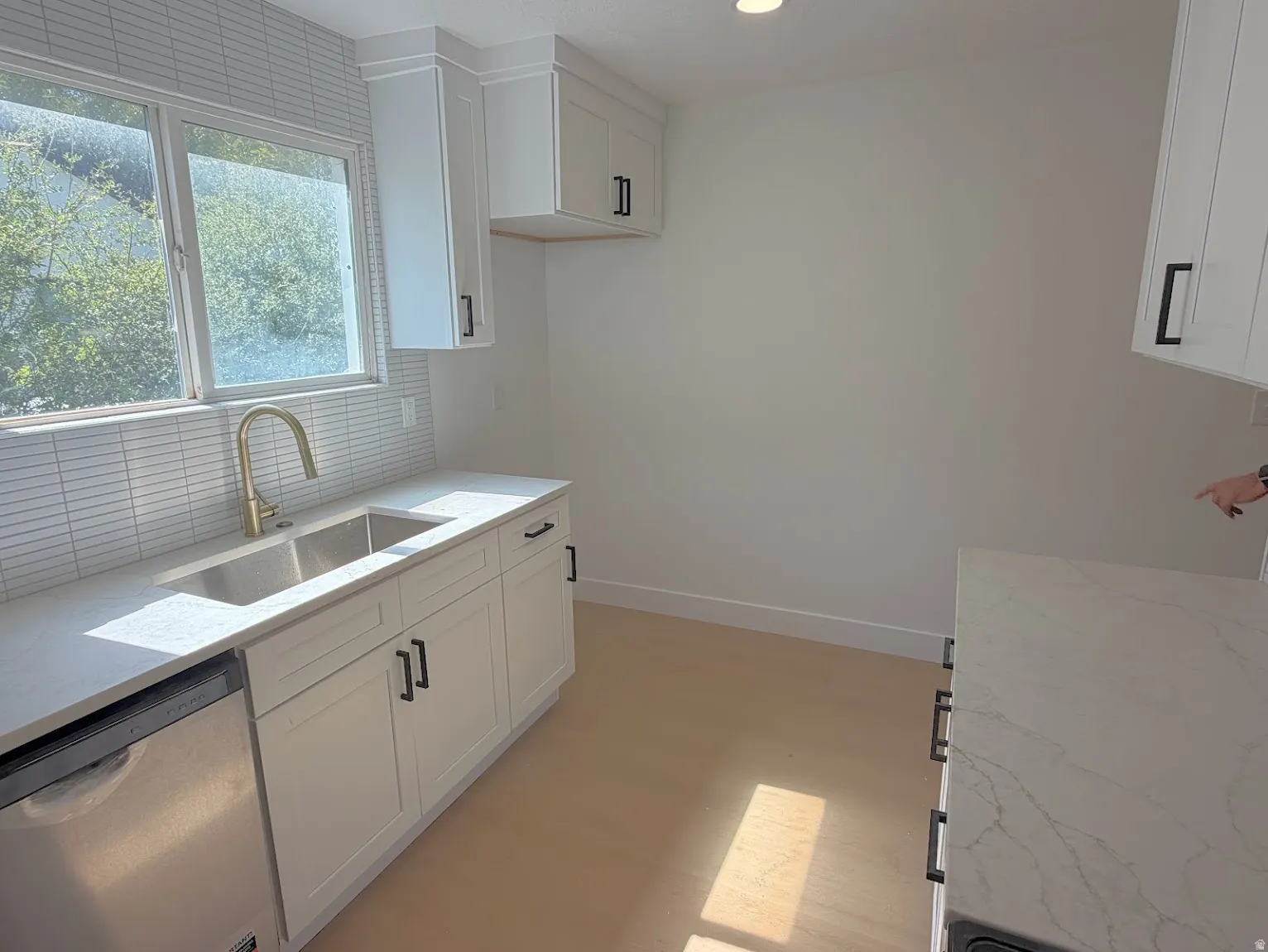White shaker-style cabinetry with black hardware, stainless steel dishwasher, double basin sink with brass finish faucet, light-toned wood-finish flooring, and white quartz countertops