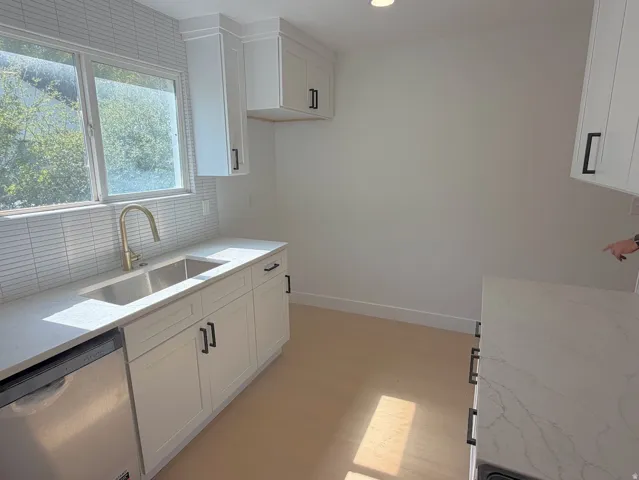 White shaker-style cabinetry with black hardware, stainless steel dishwasher, double basin sink with brass finish faucet, light-toned wood-finish flooring, and white quartz countertops
