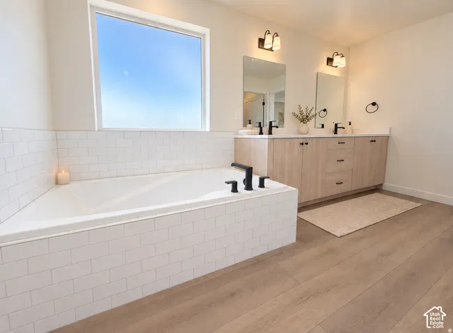 Spa-inspired primary bathroom featuring a soaking tub with matte black fixtures, dual vanities with wood-finish cabinetry and rectangular mirrors, white subway tile backsplash, warm wood-finish flooring, recessed lighting, and abundant natural light from