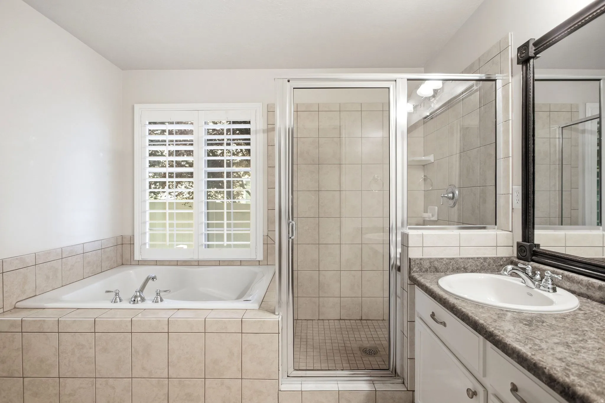 Master bathroom featuring a soaking tub with tile surround, walk-in shower with glass enclosure and tile walls, dual vanity with granite countertop and vessel sink, framed mirror, and shuttered French doors providing natural light