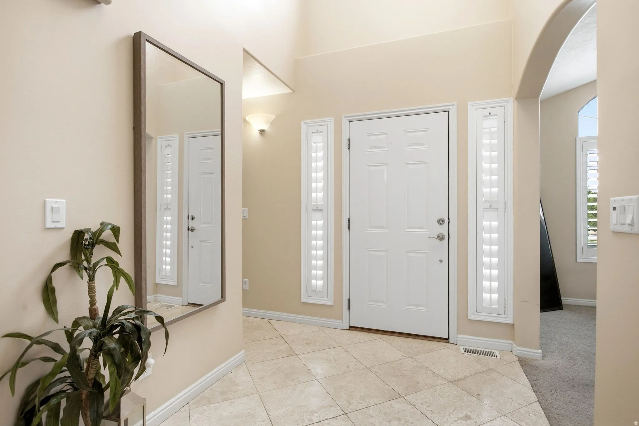 Entryway featuring tile flooring, a paneled door, two vertical window units with plantation shutters, an arched opening, and a wall-mounted sconce