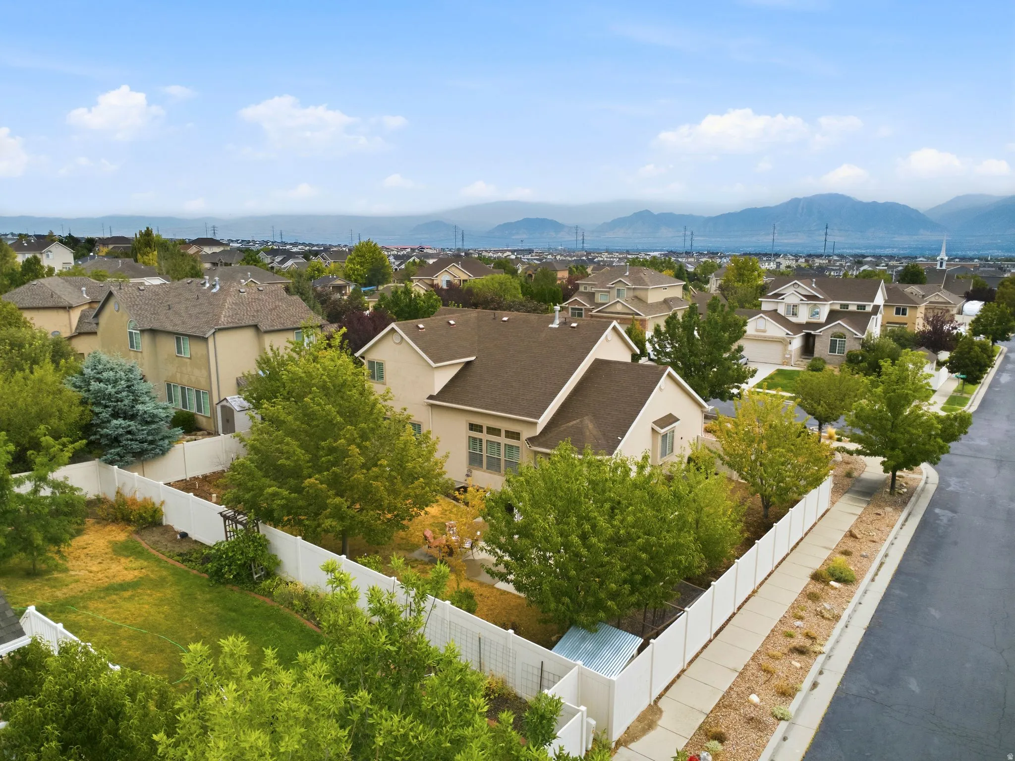 Aerial view showcasing a residence with a pitched roof and neutral exterior, situated on a corner lot with mature landscaping and white perimeter fencing