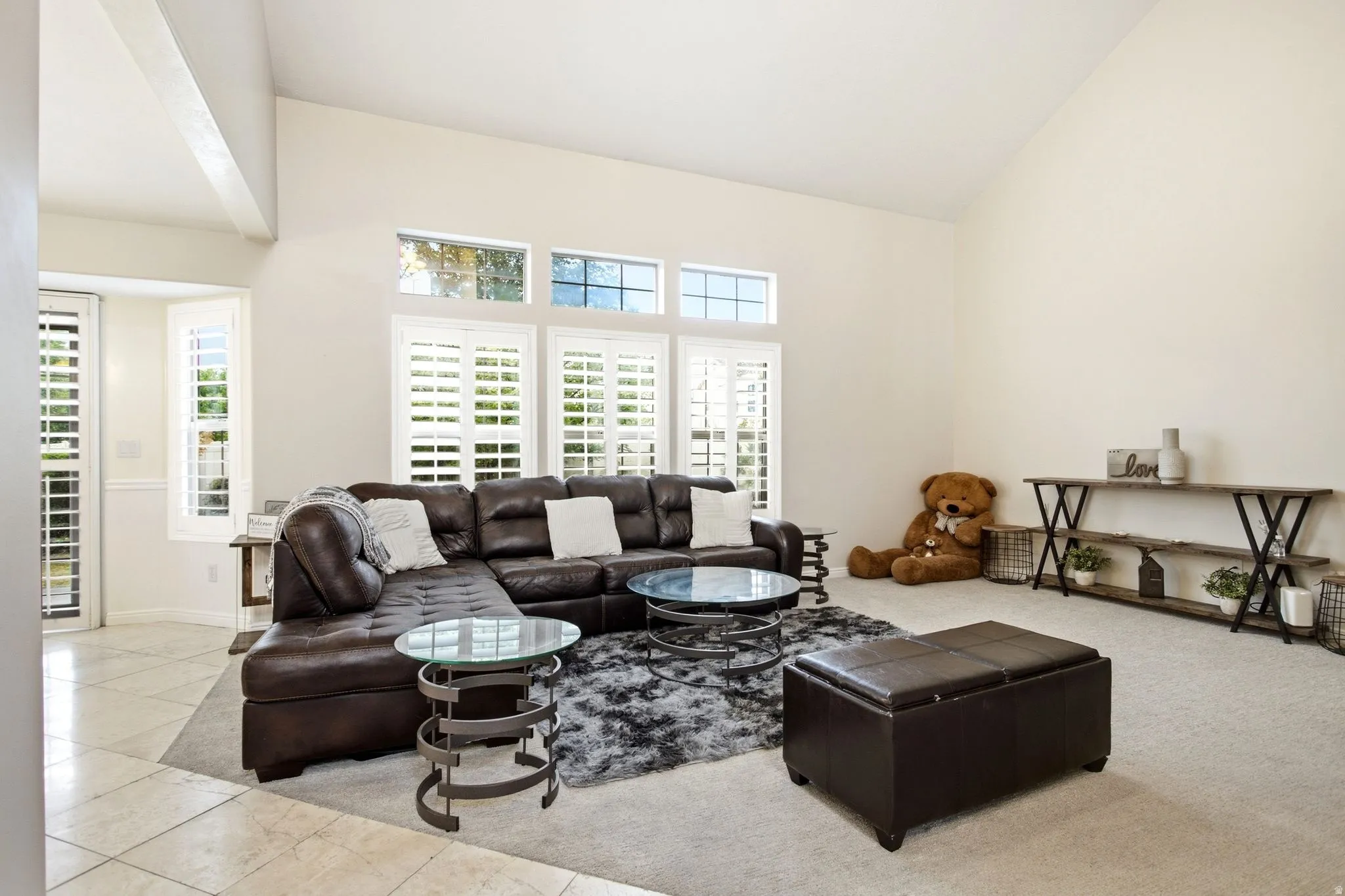 Spacious living room featuring a dark leather sectional sofa, tile flooring throughout, abundant natural light from multiple transom and shuttered windows, a round glass-top side table with metal base, and built-in shelving along the perimeter walls