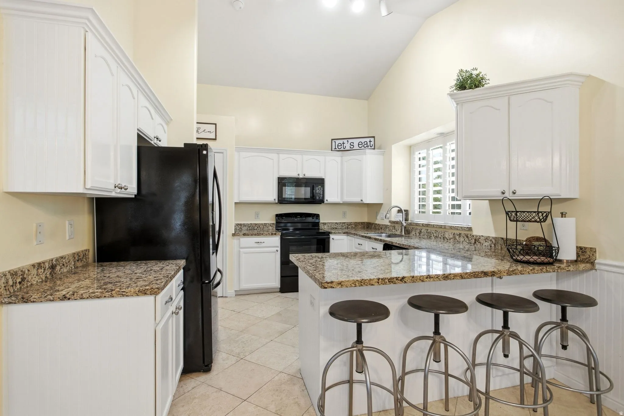 Spacious kitchen featuring white cabinetry, granite countertops, black appliances, tile flooring, and a large island with seating for multiple guests
