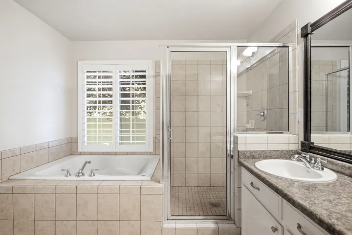 Master bathroom featuring a soaking tub with tile surround, walk-in shower with glass enclosure and tile walls, dual vanity with granite countertop and vessel sink, framed mirror, and shuttered French doors providing natural light
