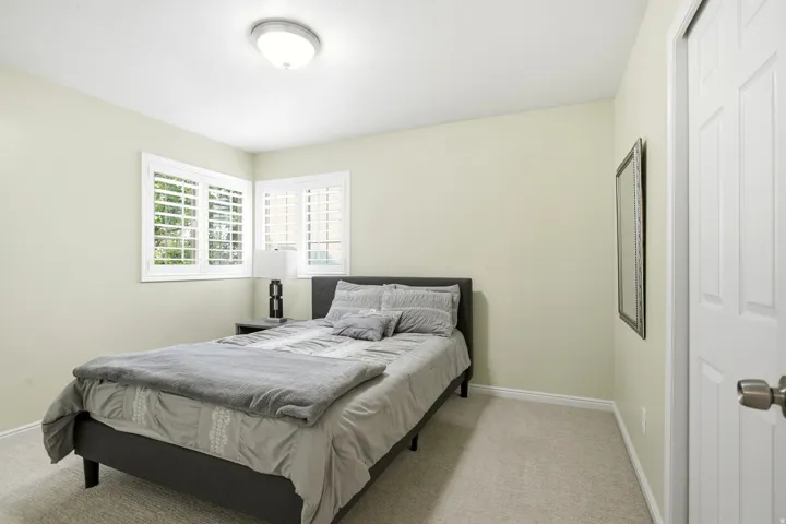 Bright secondary bedroom featuring neutral wall finishes, carpet flooring, a full-size bed frame, dual casement windows with plantation shutters, ceiling-mounted light fixture, and direct access to a full bathroom via an adjacent door