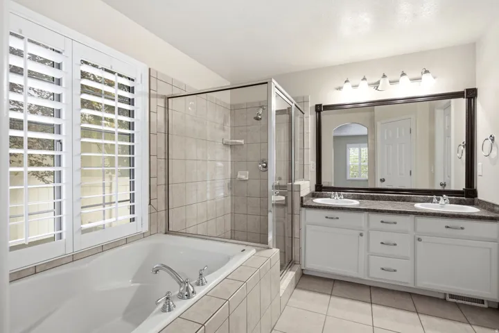 Spa-inspired primary bathroom featuring a soaking tub with tile surround, walk-in shower enclosure with glass doors, dual-sink vanity with ample storage and countertop space, framed mirror with integrated lighting, and tile flooring throughout