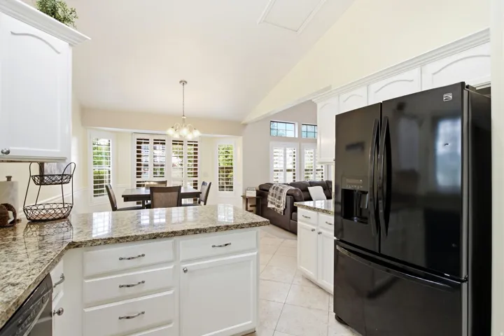 Open-concept kitchen and living area featuring white cabinetry with brushed nickel hardware, granite countertops, stainless steel French-door refrigerator, tile flooring, and abundant natural light from multiple windows with plantation shutters