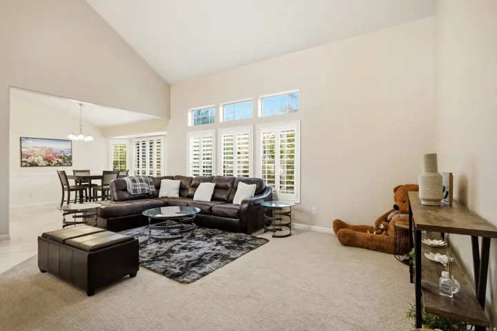 Spacious living area featuring a sectional configuration, neutral carpet flooring, and an abundance of natural light from multiple transom windows above plantation shutters