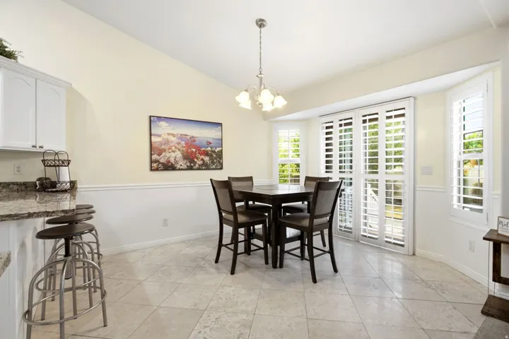 Bright and spacious dining area featuring polished stone flooring, a chandelier with multiple glass shades, and French doors with plantation shutters opening to outdoor space