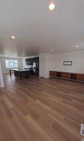 Spacious open-concept living area featuring wood-finish flooring, recessed lighting, a modern kitchen with dark cabinetry and stainless steel appliances, and a built-in media console
