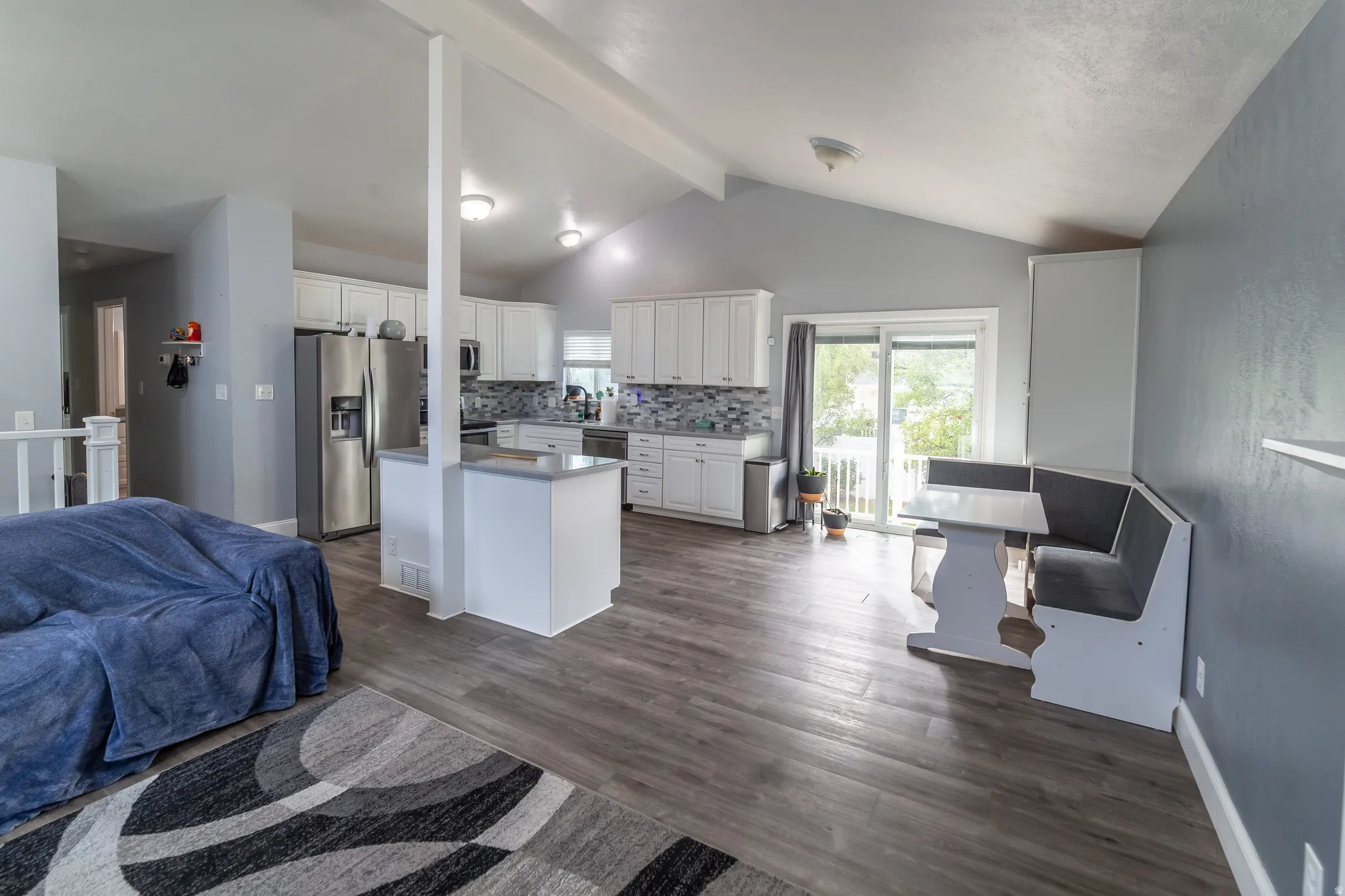 Open-concept living space featuring vaulted ceilings, wood-finish flooring, a kitchen island with stone countertop, white cabinetry, and stainless steel appliances