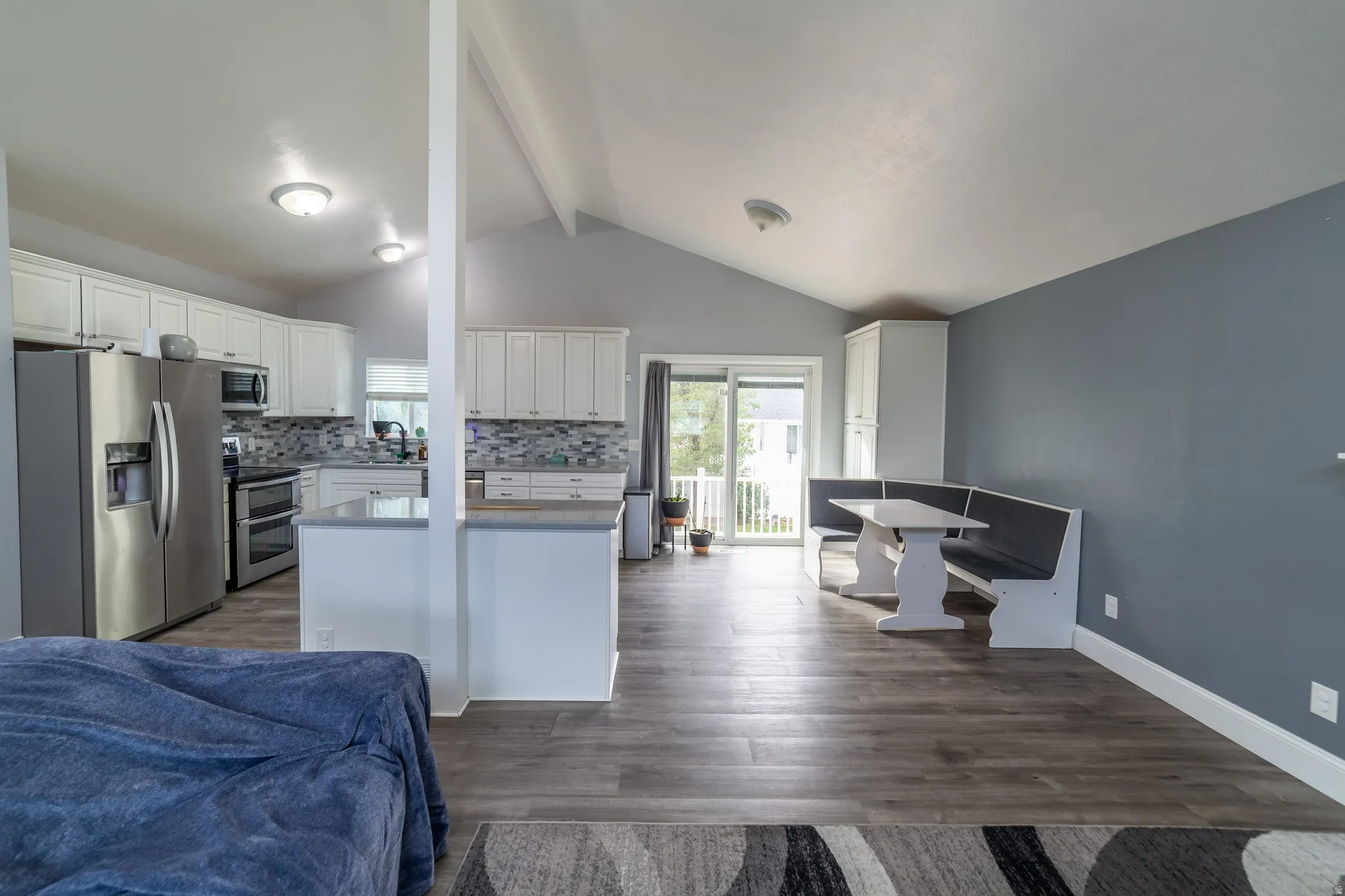 Vaulted ceiling, wood-finish flooring, kitchen with white cabinetry, stainless steel appliances, and a tiled backsplash, and a sliding glass door