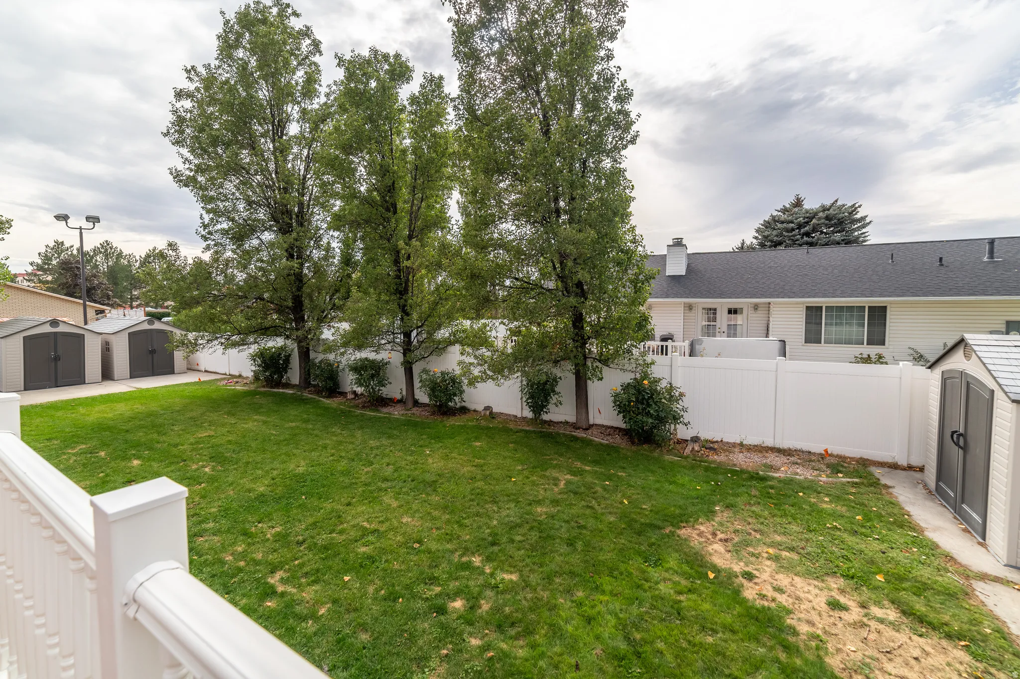 Expansive yard with green lawn, mature trees, white privacy fencing, and two storage sheds