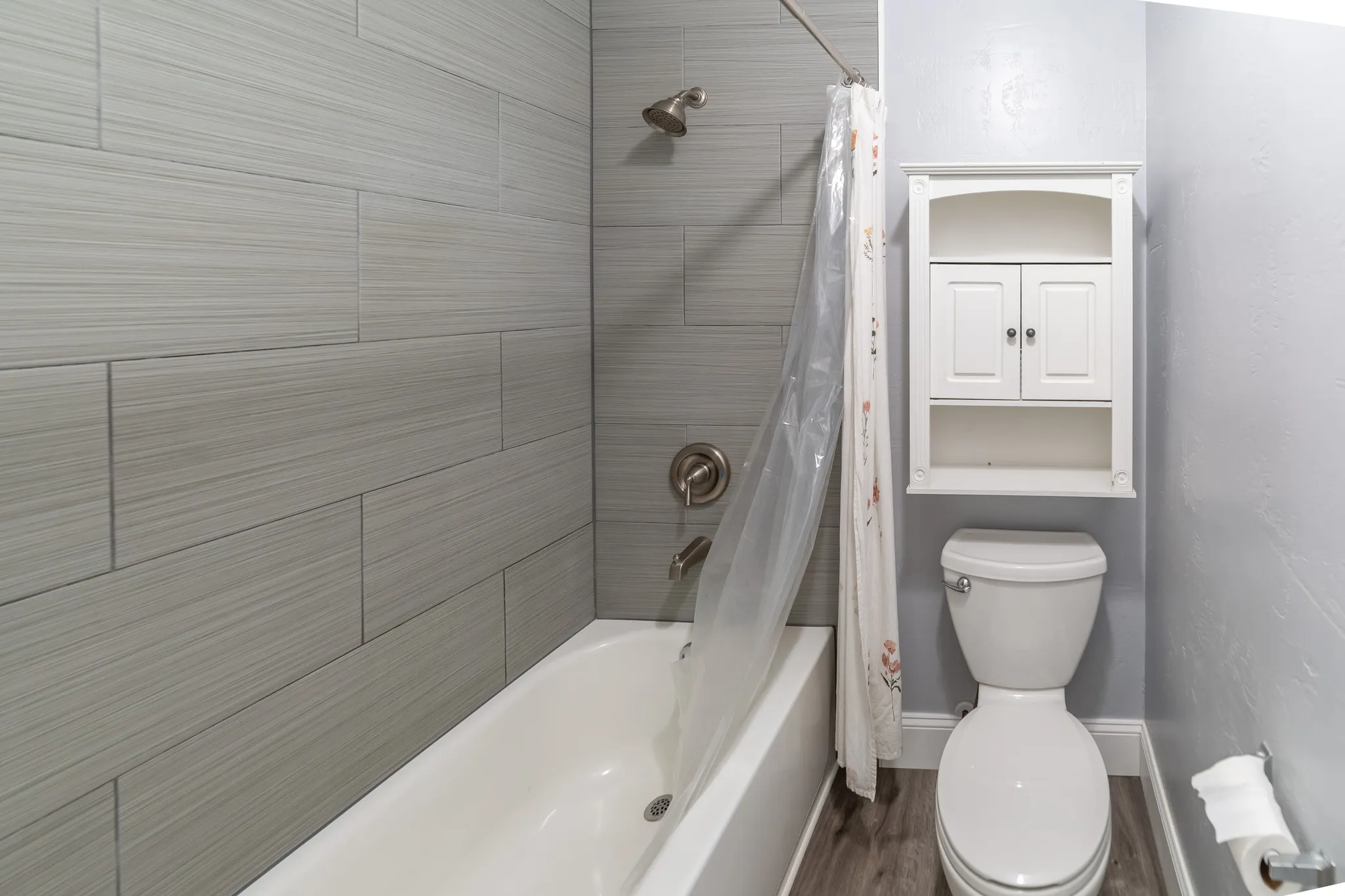 Bathtub with brushed nickel fixtures, light gray wall tiling, wood-finish flooring, and a wall-mounted storage cabinet