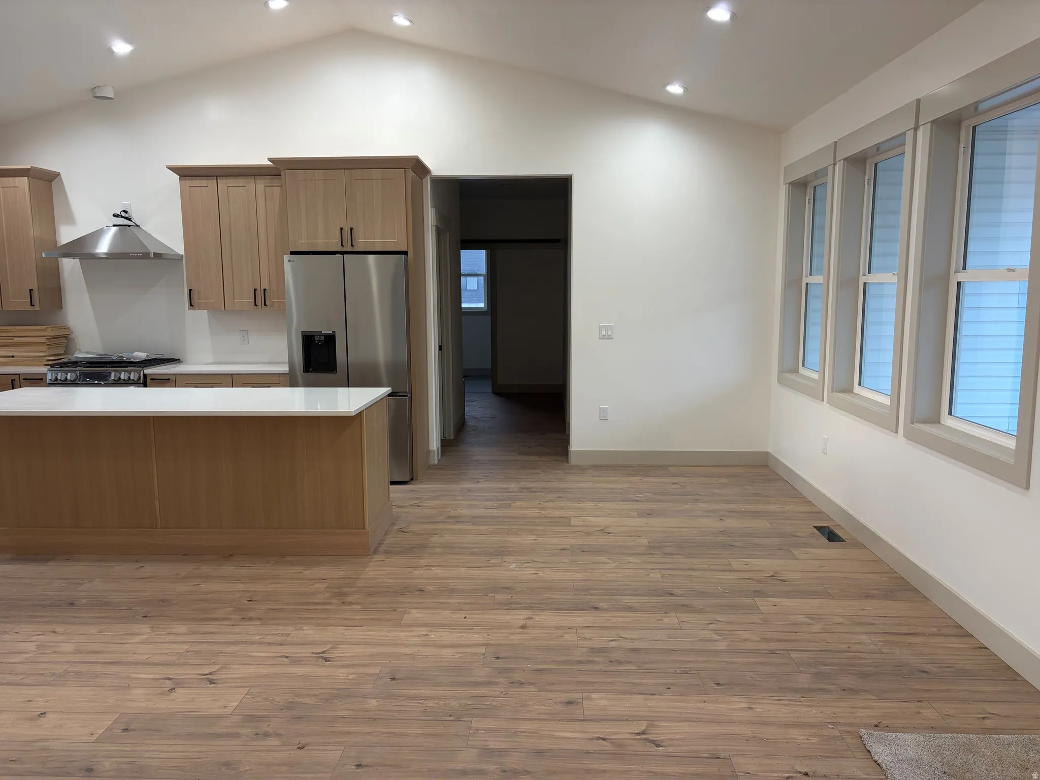 Vaulted ceiling with recessed lighting, wood-finish flooring, kitchen island with a white countertop, stainless steel appliances, and multiple windows