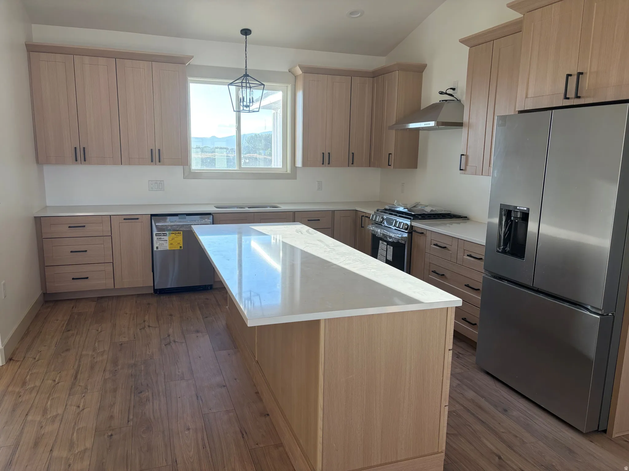 Kitchen featuring light wood-finish cabinetry, stainless steel appliances, a center island with a light-toned countertop, wood-finish flooring, and a contemporary pendant light fixture
