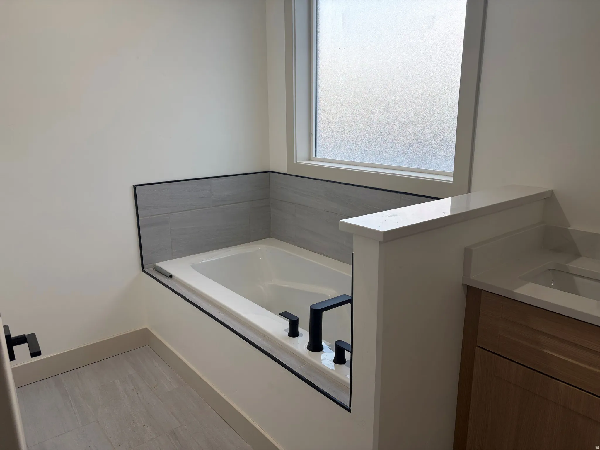 Built-in soaker tub with matte black fixtures, gray wall tiling, and privacy window