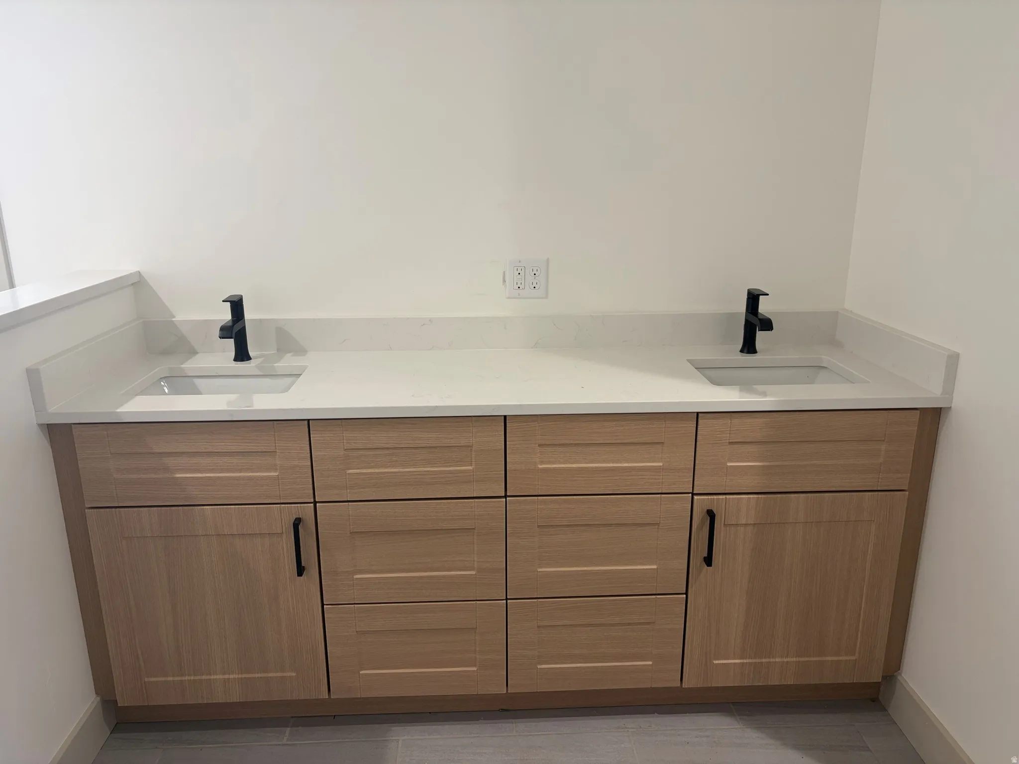 Dual vanity with light wood-finish cabinetry, integrated sinks, black fixtures, and light-toned countertops