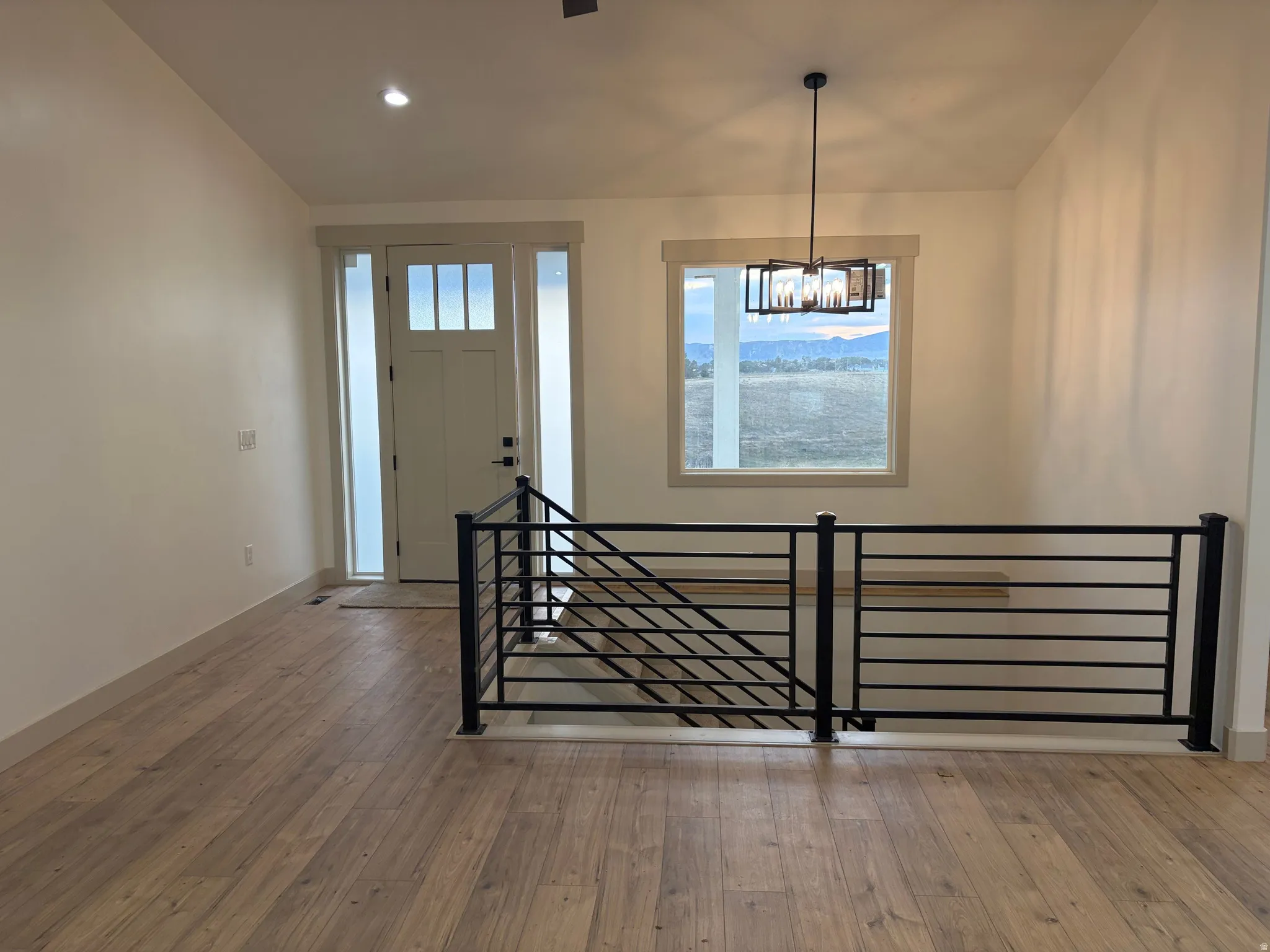 Wood-finish flooring, contemporary black metal railing, prominent window with outdoor scenery, modern entry door with sidelights, and a decorative overhead light fixture
