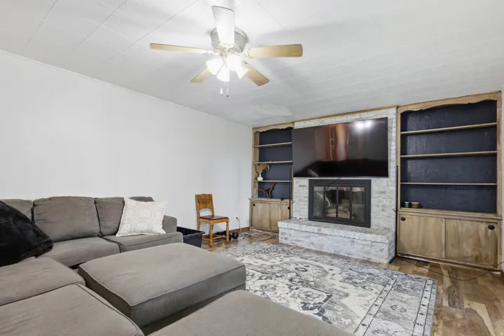 Living area featuring wood-finish flooring, a ceiling fan, and a fireplace with a light-colored brick surround