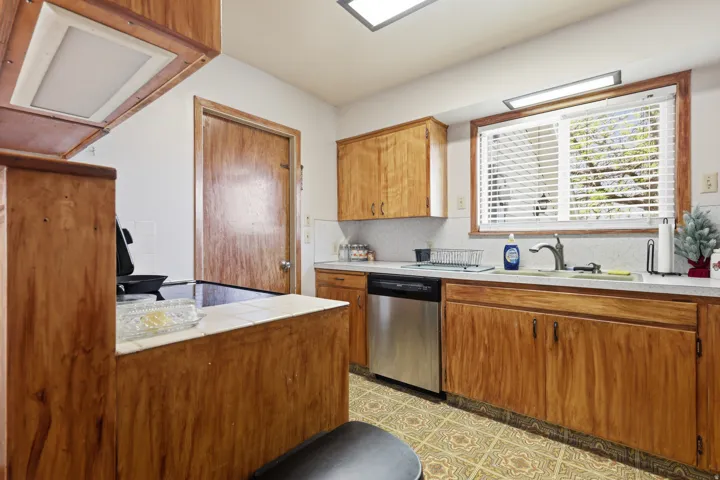Wood-finish cabinetry, tile countertops, stainless steel dishwasher, tile flooring, and a kitchen window