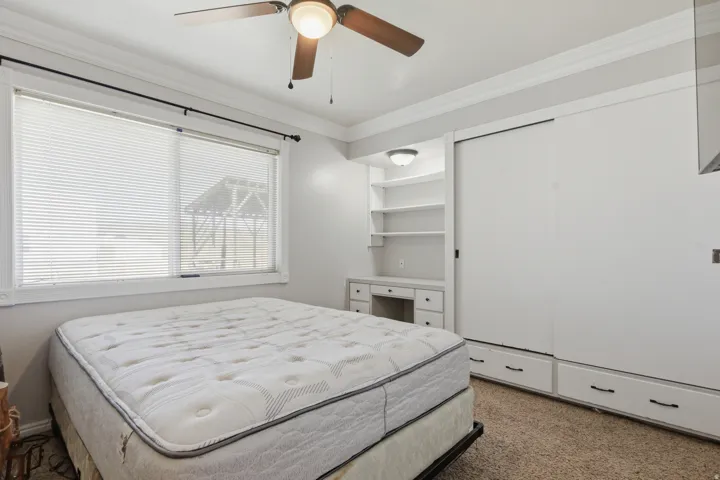 Ceiling fan with light fixture, large window with blinds, built-in shelving, built-in desk, and built-in closet storage