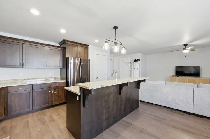 Kitchen island with integrated sink, dark wood-finish cabinetry, light-toned countertops, stainless steel refrigerator, and wood-finish flooring