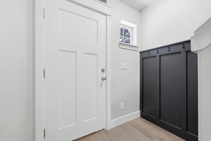 White paneled door, built-in black mudroom bench with hooks, wood-finish flooring, and natural light from a transom window