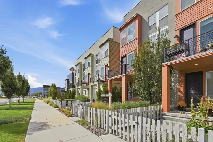 Contemporary townhomes featuring varied exterior finishes, individual balconies with black railings, a white picket fence, and concrete sidewalks