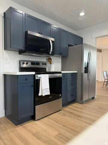 Kitchen featuring dark shaker-style cabinetry, stainless steel appliances, light countertops, and wood-finish flooring