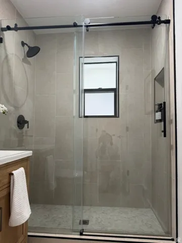 Glass-enclosed shower with a black-framed window, large-format wall tiles, and a pebble-finish shower floor