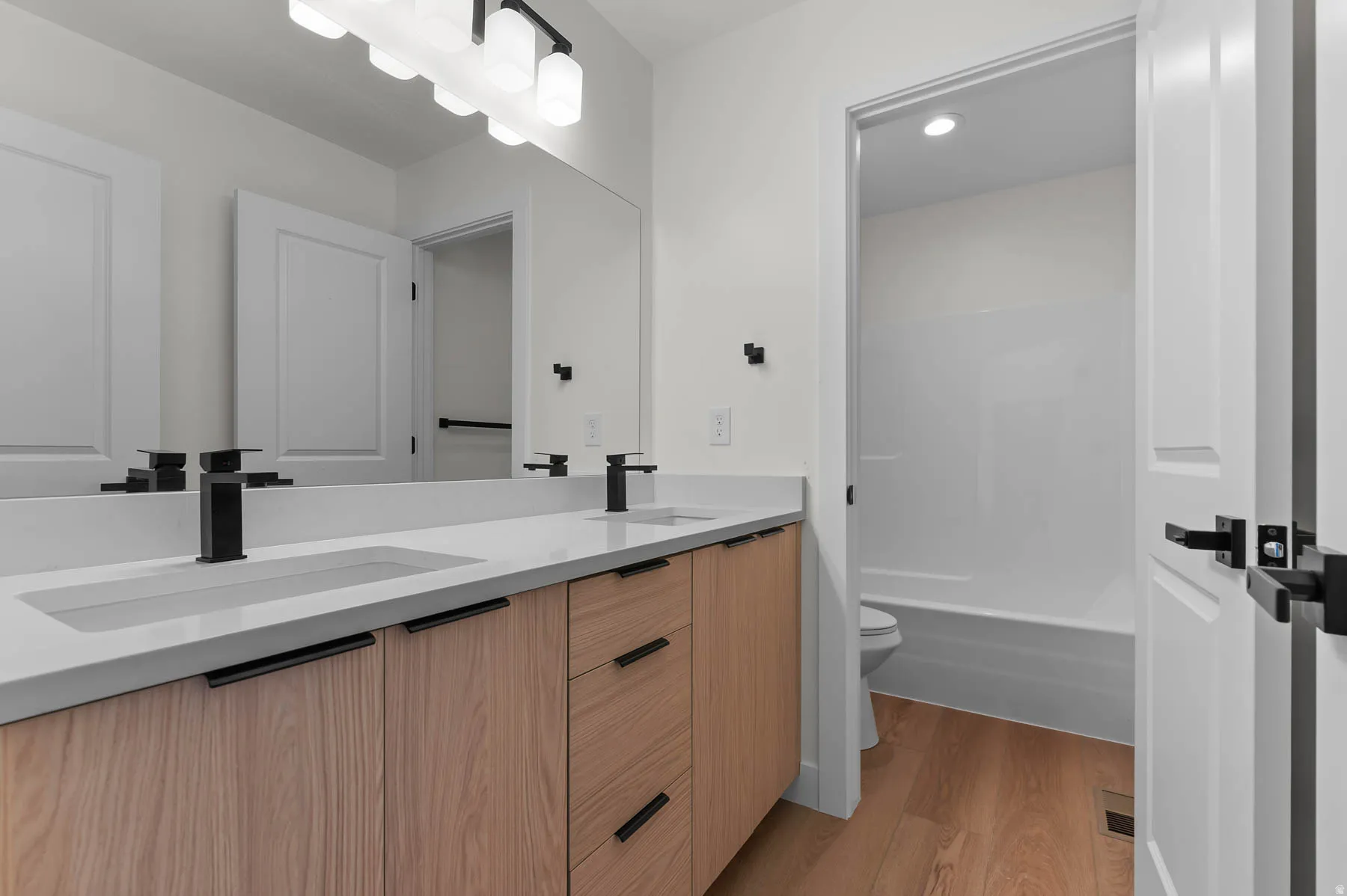 Dual undermount sinks with matte black faucets, wood-finish vanity with drawer storage, large wall-mounted mirror, and overhead vanity lighting