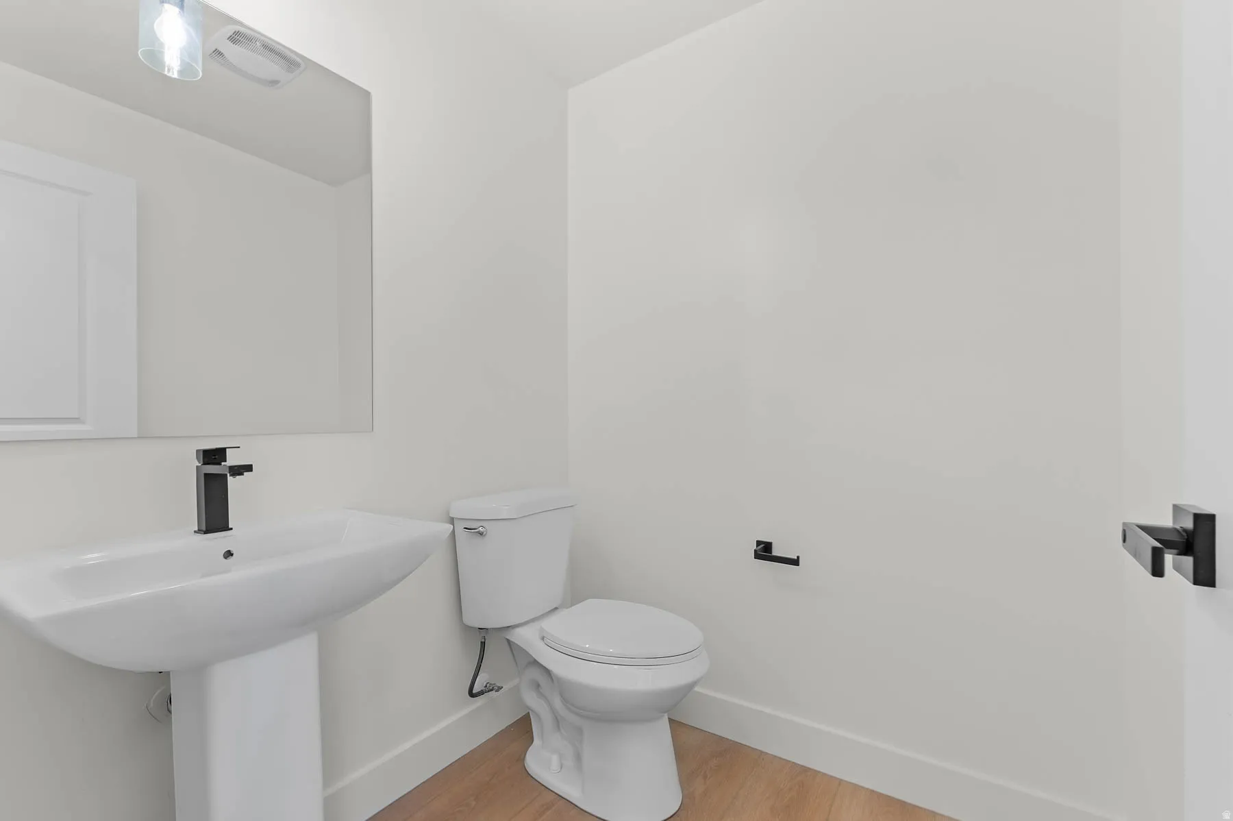 Pedestal sink with a matte black faucet, wall-mounted mirror, and wood-finish flooring