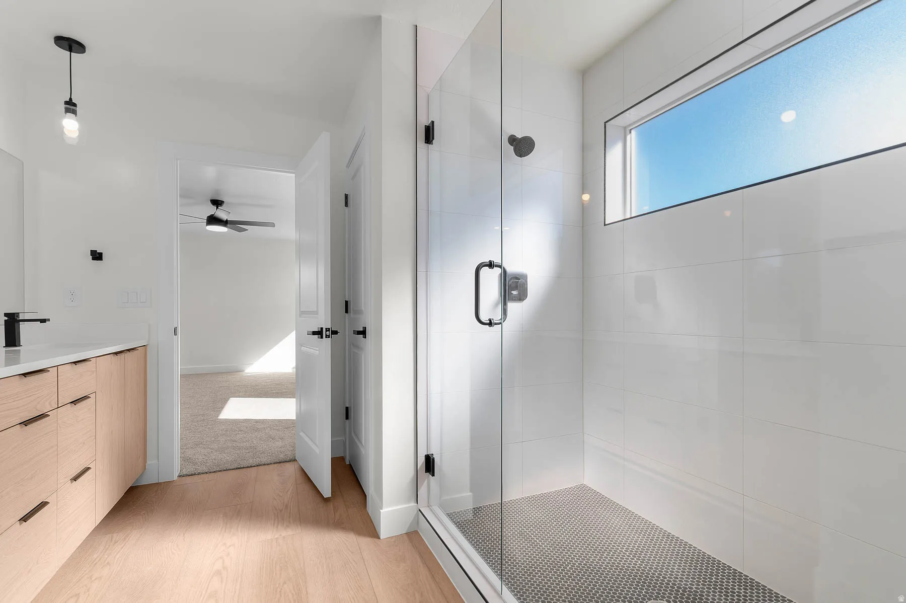 Glass-enclosed shower with a mosaic tile floor, wide plank wood-finish flooring, a wood-finish vanity with a white countertop, and a rectangular transom window