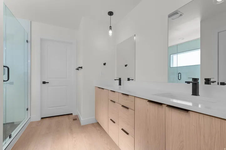 Dual vanity with integrated sinks, light wood-finish cabinetry, matte black fixtures, and a clear glass enclosed shower