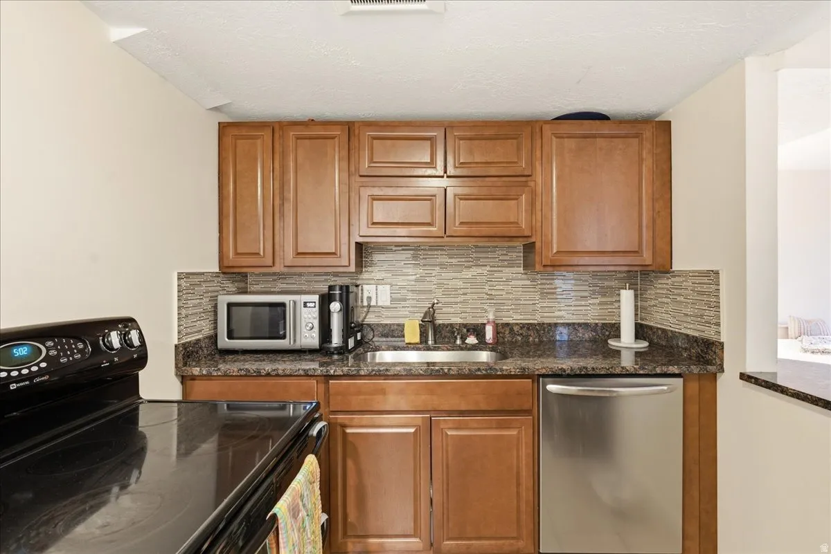 Wood-finish cabinetry, dark speckled countertops, tile backsplash, and a stainless steel dishwasher