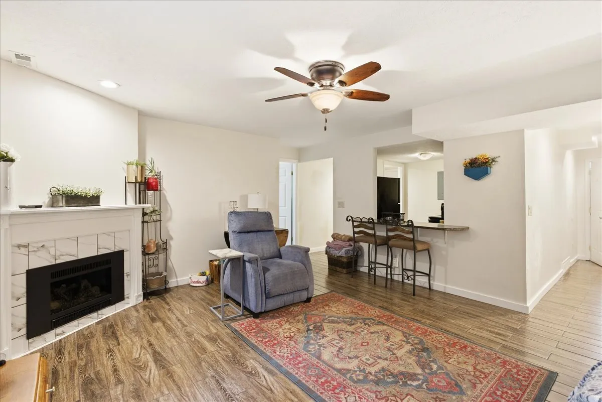 Wood-finish flooring throughout, white fireplace with tile surround, ceiling fan with light fixture, recessed lighting, and a built-in bar counter