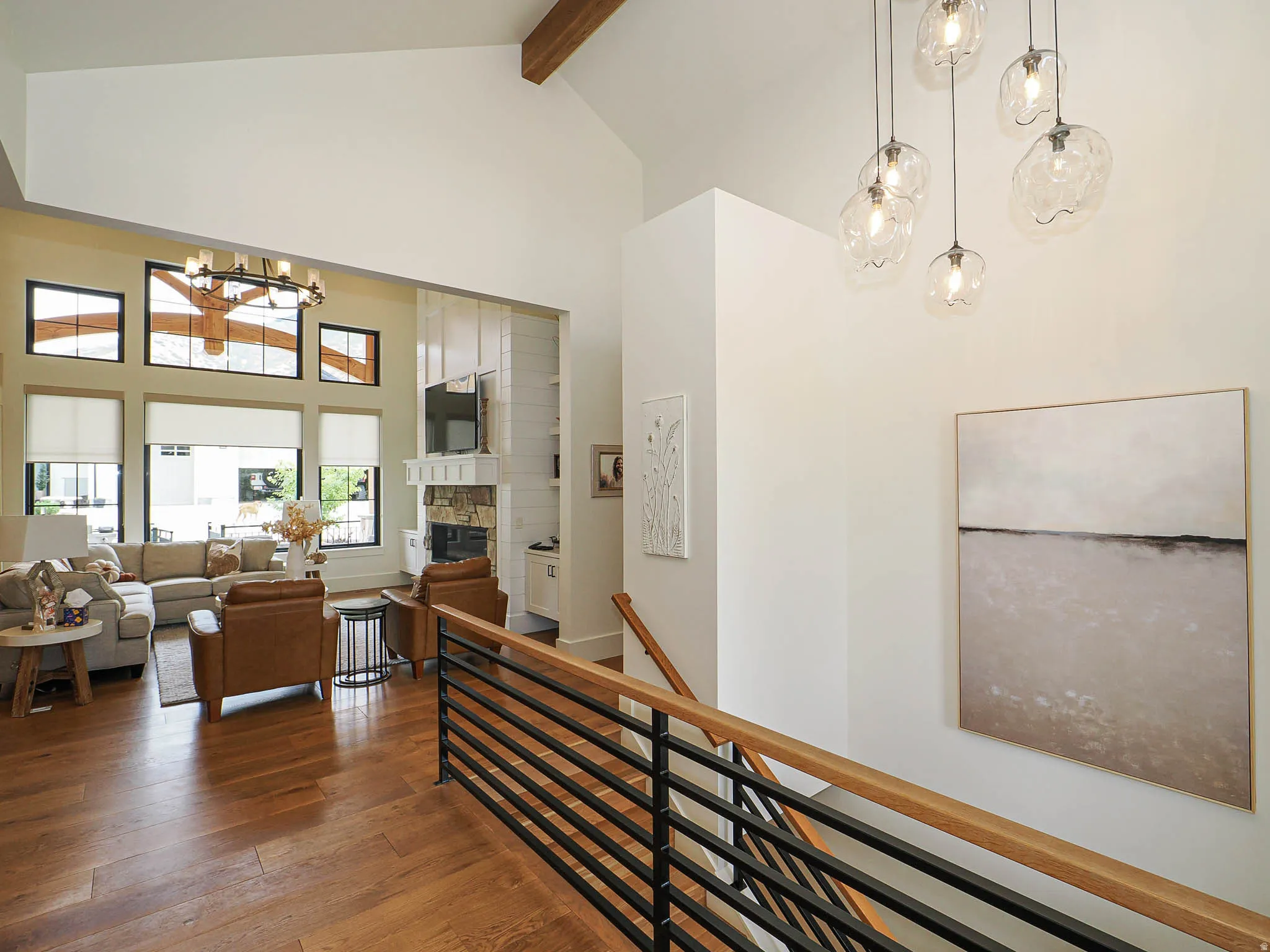 Vaulted ceiling with exposed wood beam, wood-finish flooring, staircase with wood handrail and black metal balusters, stacked stone fireplace with built-in shiplap cabinetry, and multiple large windows