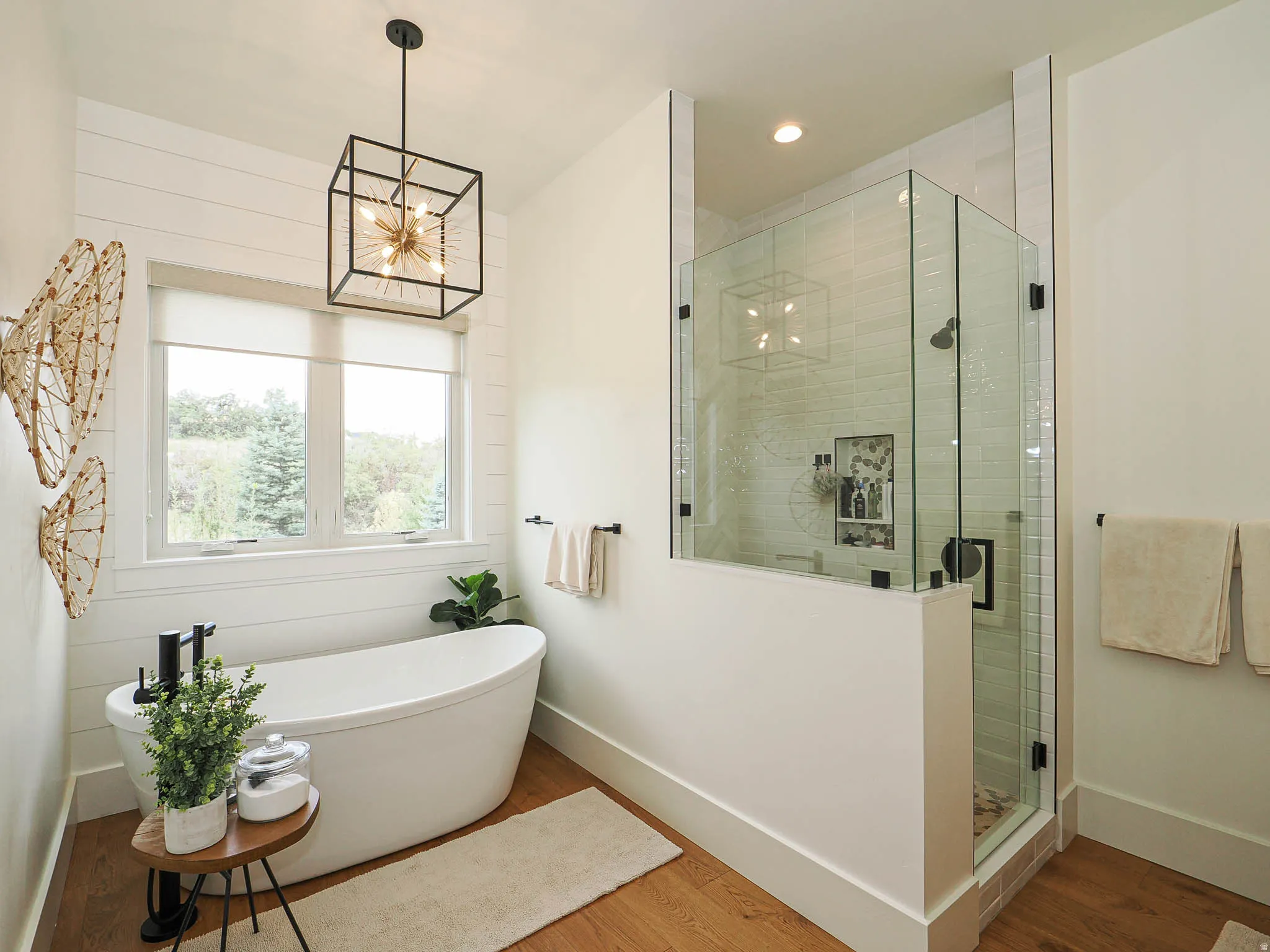 Freestanding soaking tub with matte black fixtures, glass-enclosed shower with tile surround, wood-finish flooring, and white shiplap wall accent