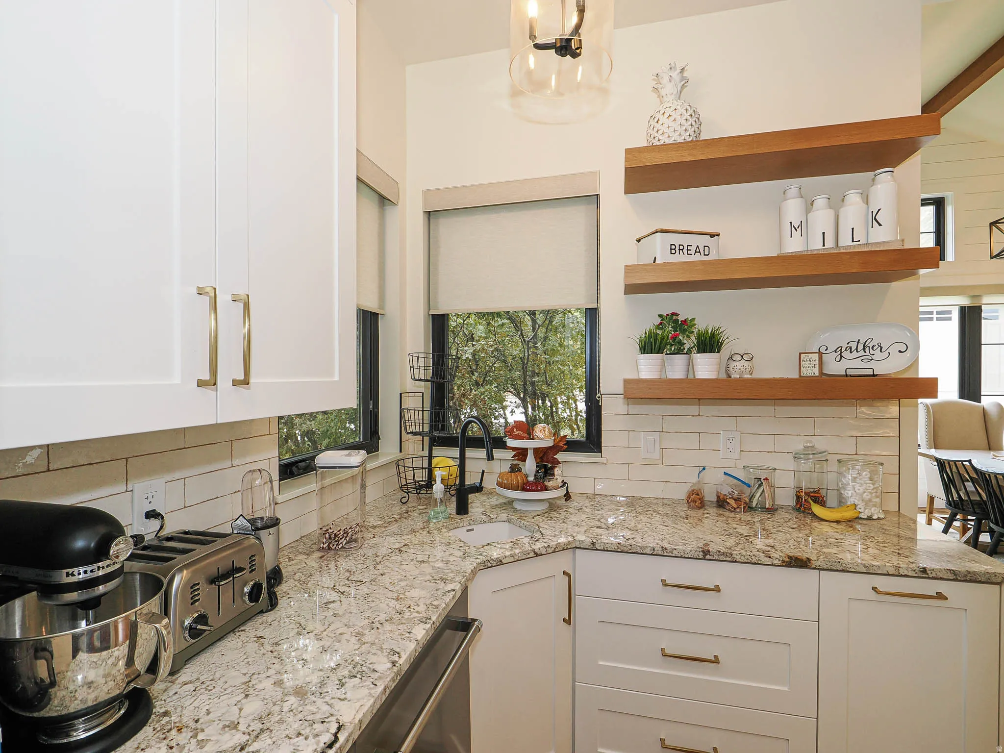 White shaker cabinetry with gold-tone hardware, granite countertops, subway tile backsplash, and floating wood shelving