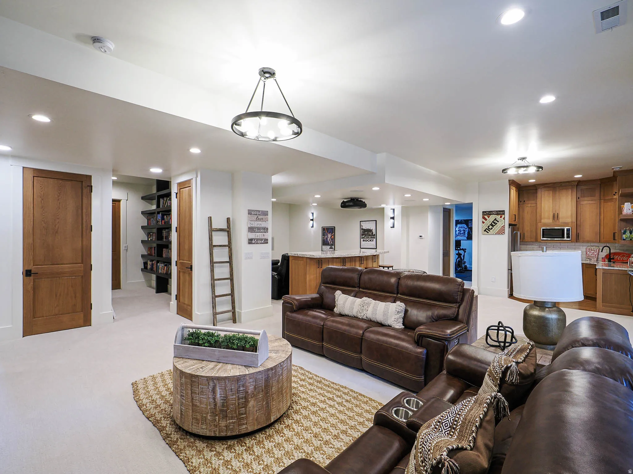 Recessed lighting and a decorative chandelier illuminate a spacious room featuring a built-in entertainment bar, warm wood cabinetry, and a dedicated shelving alcove