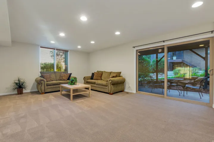 Recessed ceiling lighting, full-height sliding glass doors, window with horizontal blinds, neutral carpet flooring, and a patio with outdoor seating