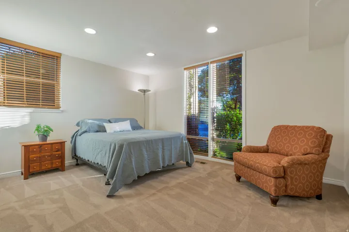 Recessed ceiling lighting illuminates the carpeted room, featuring a large window with horizontal blinds and an additional window with horizontal blinds